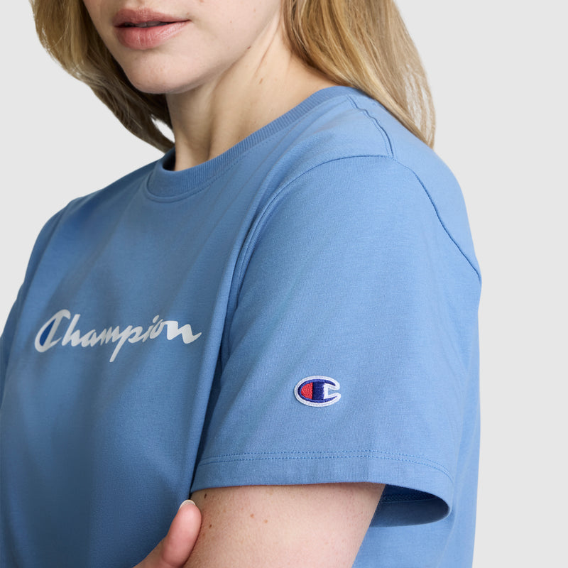 A person wears a light blue Champion Classic T-Shirt, featuring the script logo on the chest and sleeve, against a plain background.
