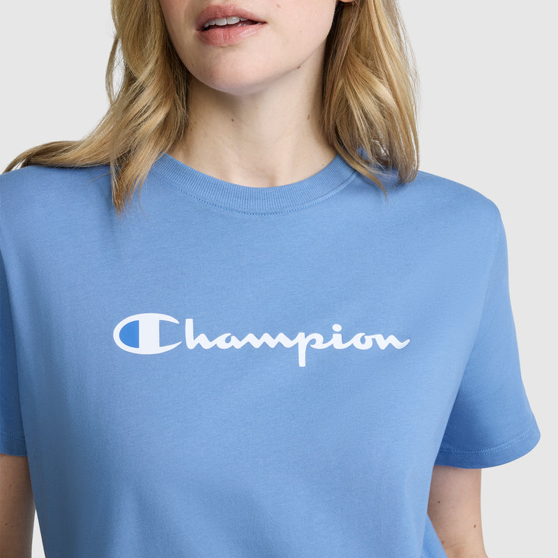 A woman with light brown hair is shown from the shoulders to the mouth against a plain background, wearing a light blue Champion Classic T-Shirt with the signature script logo.