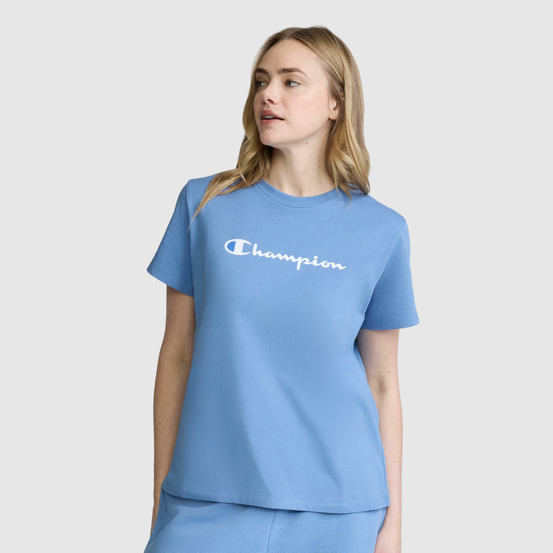 A woman stands against a plain white background, looking to her left, dressed in a light blue Champion Classic T-Shirt with the signature script logo and matching shorts.
