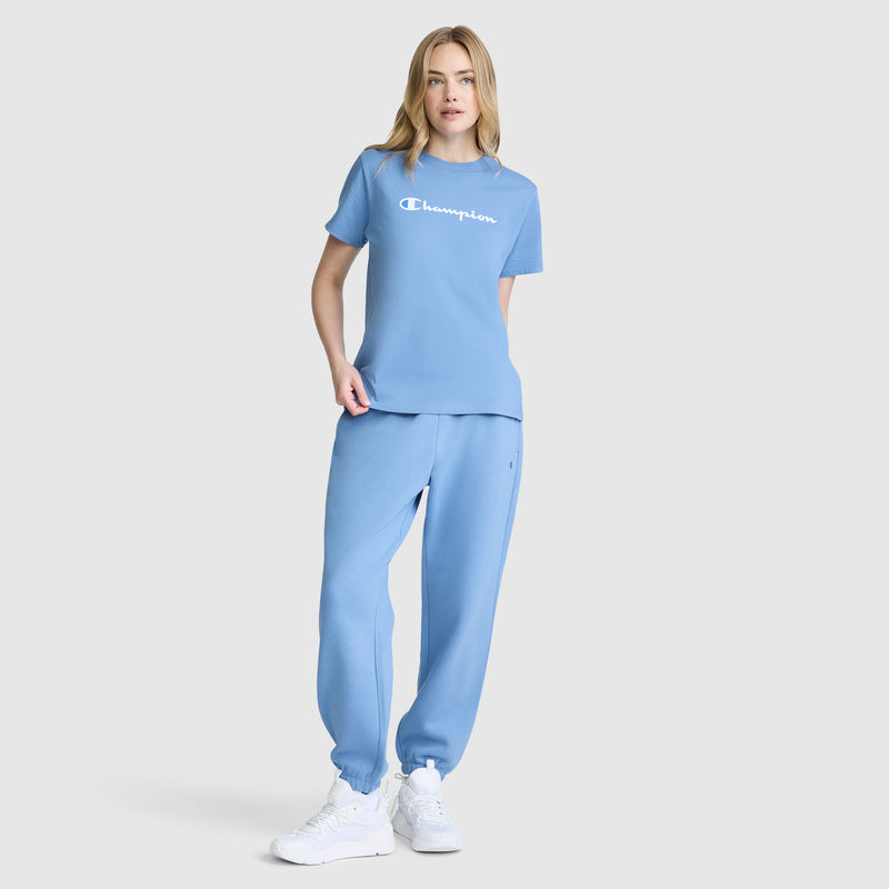 A woman stands against a plain background, dressed in a light blue Champion Classic T-Shirt with the signature script logo, paired with matching sweatpants and white sneakers.