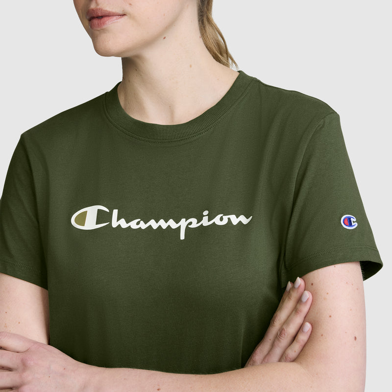 A woman stands with her arms crossed, wearing the Champion Classic T-Shirt, Script Logo in olive green. This women’s athletic shirt showcases the signature white script logo across the chest and a small Champion patch on the left sleeve, combining comfort and iconic style.