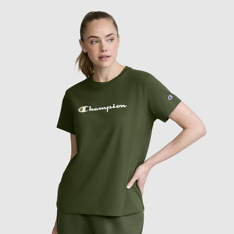 A woman wearing an olive green Champion Classic T-Shirt with the script logo and matching pants stands with one hand on her hip, gazing slightly to the side against a light gray background.