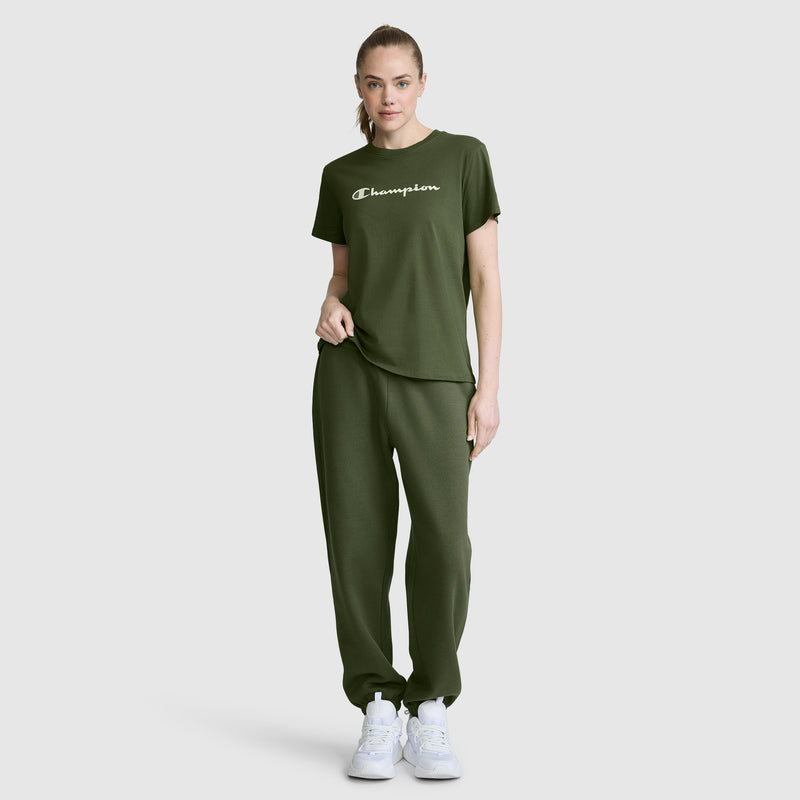A woman stands against a plain light gray background wearing an olive green Champion Classic T-Shirt with the script logo and matching sweatpants, paired with white sneakers.