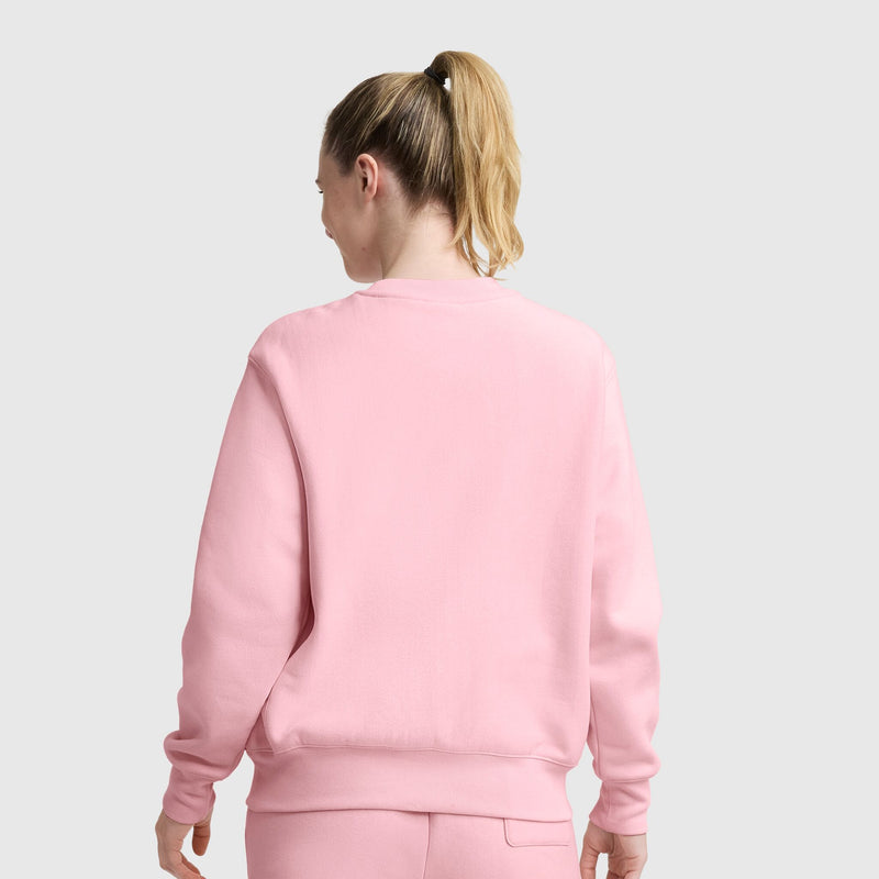 A woman with blonde hair in a ponytail stands facing away from the camera, wearing a light pink Champion Essentials Reverse Weave Crew Neck Sweatshirt and matching pants, exuding timeless comfort against a plain light gray background.