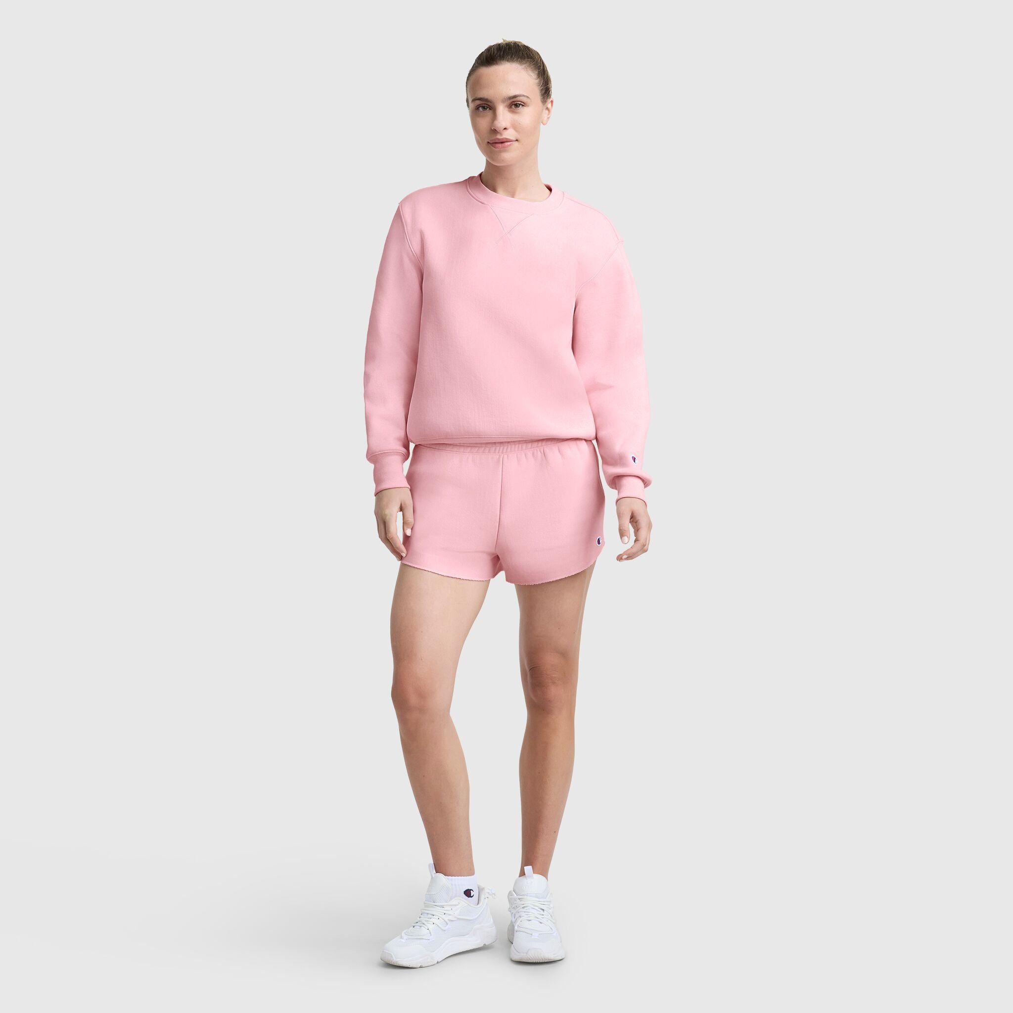 A woman stands against a light gray background, wearing the Champion Essentials Reverse Weave Crew Neck Sweatshirt and matching shorts in light pink with white sneakers, looking ahead with a neutral expression and relaxed posture.