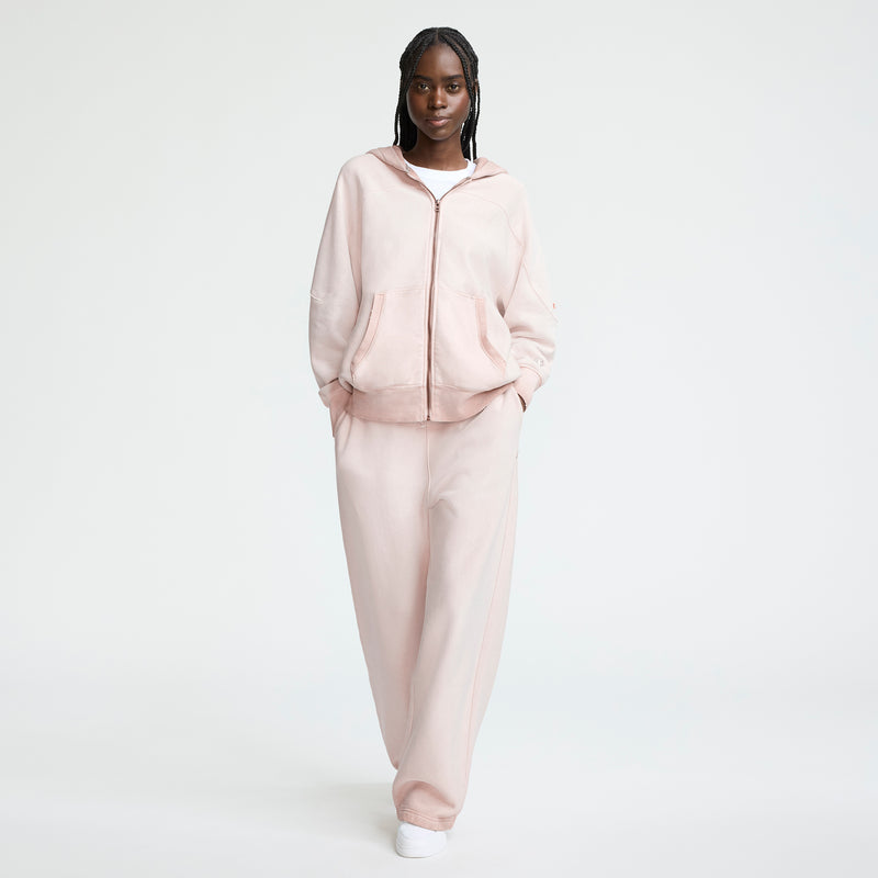 A woman stands against a plain white background wearing a Champion Time Capsule Vintage 80's Full Zip Hoodie in light pink, matching loose sweatpants, and white sneakers, with her hands in her pockets.