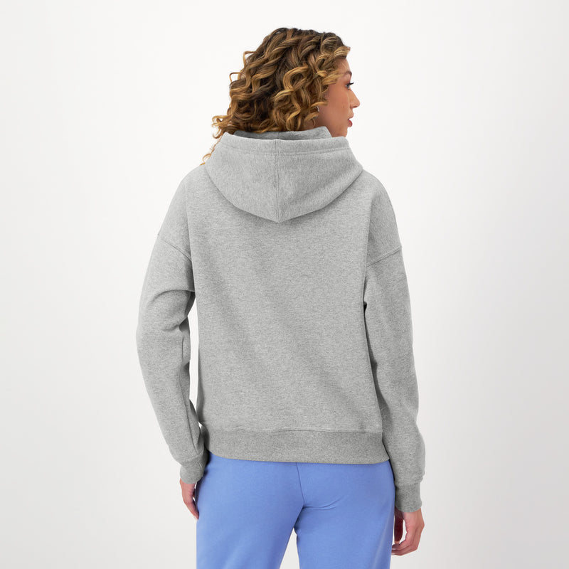 A person with curly hair is seen from the back in a light gray Champion Powerblend Freshman Hoodie with embroidered script logo and light blue pants, standing against a plain white background.