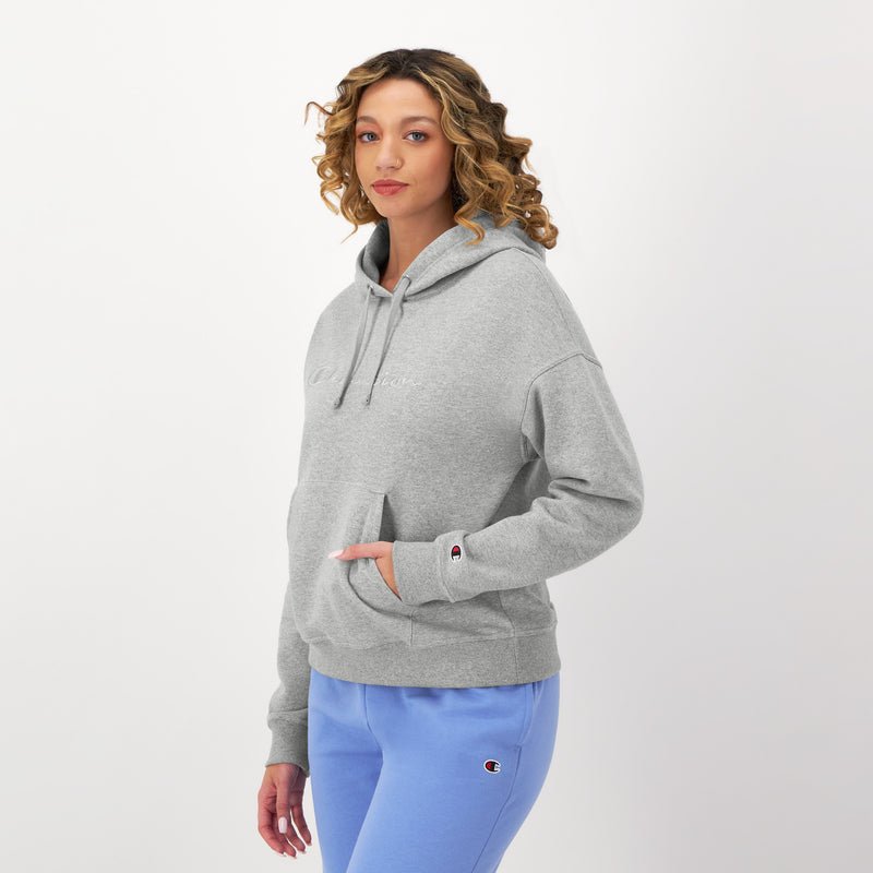 A woman with curly hair wears a light gray Champion Powerblend Freshman Hoodie with an embroidered script logo and light blue sweatpants, both displaying Champion logos, standing against a plain white background with one hand in her hoodie pocket.
