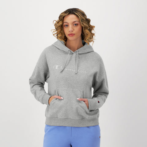 A woman with curly hair stands against a white background, hands in the front pocket of a light gray Champion Powerblend Freshman Hoodie with an embroidered script logo and wearing light blue sweatpants.