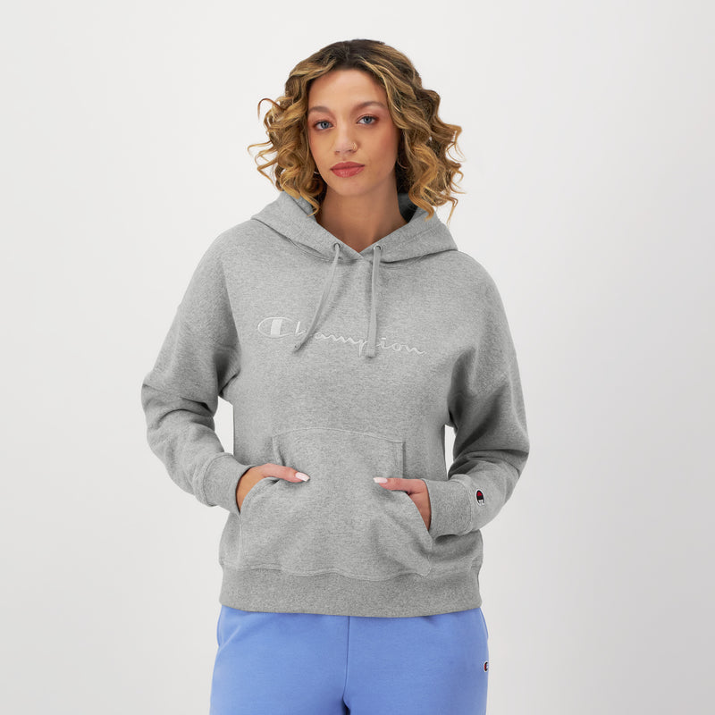 A woman with curly hair stands against a white background, hands in the front pocket of a light gray Champion Powerblend Freshman Hoodie with an embroidered script logo and wearing light blue sweatpants.
