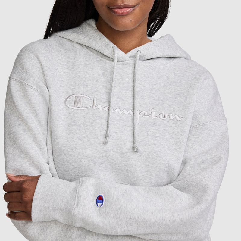 A woman wearing a light gray Champion Tonal Script Freshman Powerblend Hoodie, a cozy fleece hoodie featuring an embroidered logo on the chest and a small logo patch on the sleeve, stands with her arms crossed. This classic women's sweatshirt from Champion effortlessly combines comfort and style for any casual occasion.
