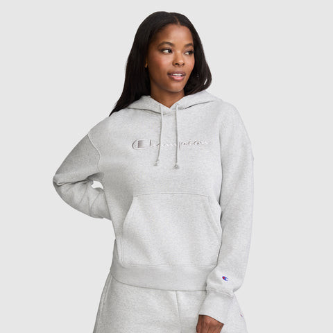 A woman wearing a light gray Champion Tonal Script Freshman Powerblend Hoodie and matching sweatpants stands against a plain light gray background, looking slightly to her left with one hand on her hip. This cozy fleece Tonal Script Freshman Powerblend Hoodie from Champion offers both comfort and style, making it the perfect women's hooded sweatshirt for everyday wear.