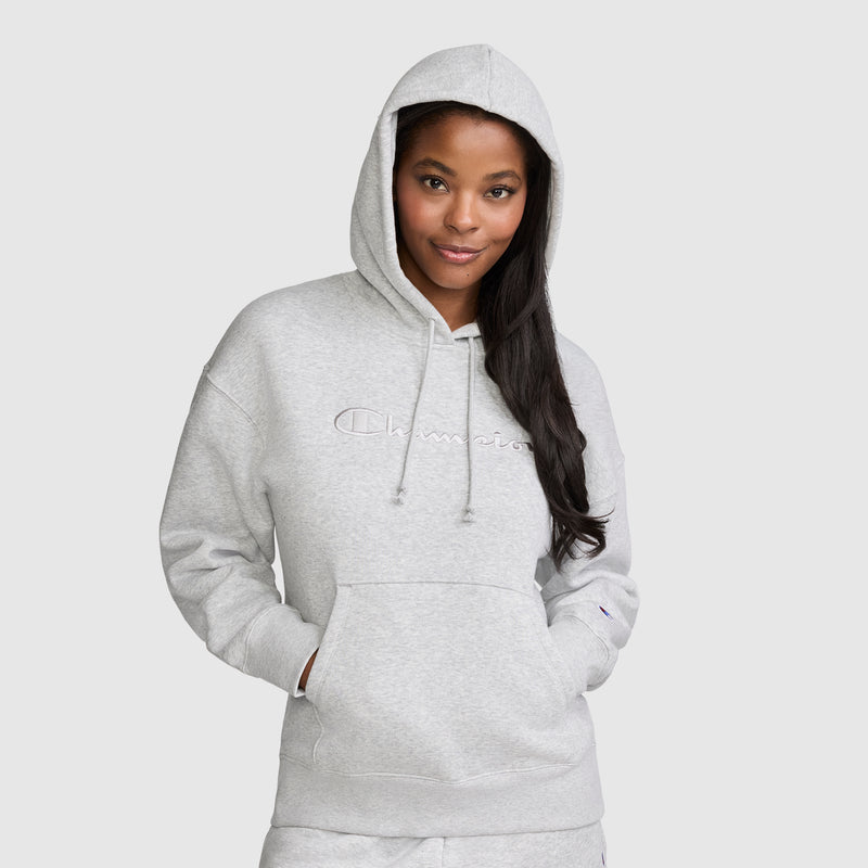 A woman wearing a light gray Champion Tonal Script Freshman Powerblend Hoodie with "Champion" embroidered on the front stands with her hands in the front pocket, smiling slightly against a plain, light background. This cozy hoodie is crafted from soft fleece, providing both comfort and style.