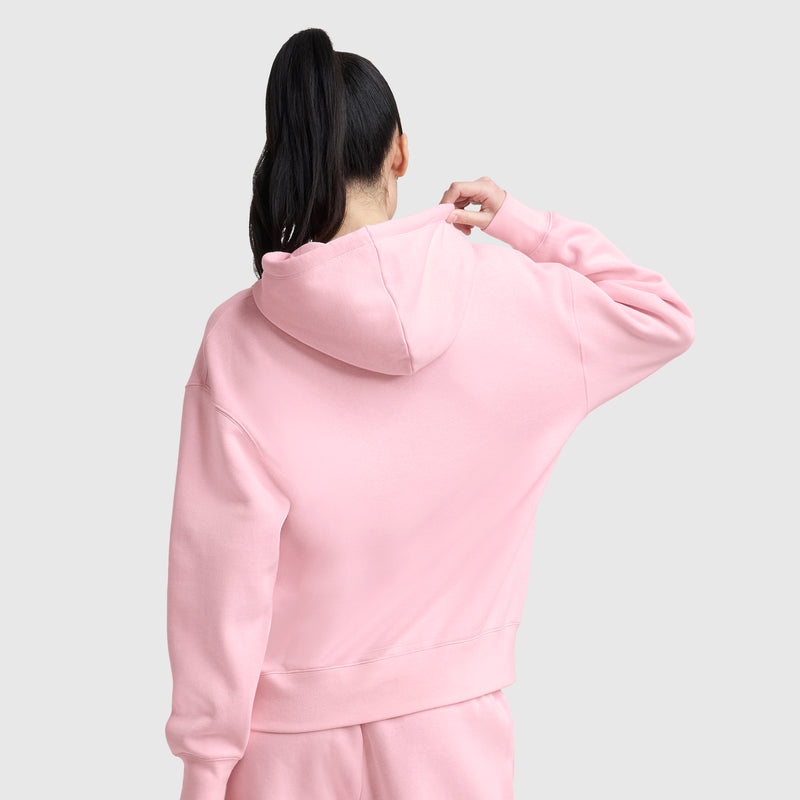 A person with long dark hair in a ponytail is shown from behind, wearing the Champion Tonal Script Freshman Powerblend Hoodie and matching pants in light pink. They hold the hood with one hand against a plain light gray background.