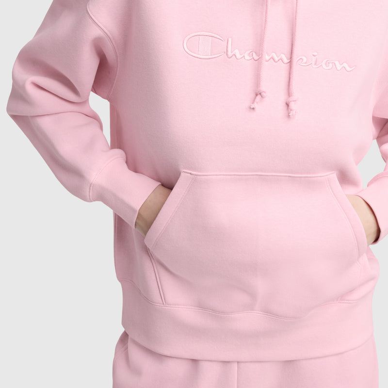 A person wears a light pink Champion Tonal Script Freshman Powerblend Hoodie with matching sweatpants, hands in the front pocket, shown from shoulders to hips against a plain light background.