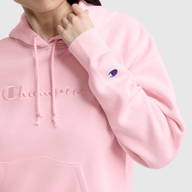 A person wearing a light pink Champion Tonal Script Freshman Powerblend Hoodie, featuring the brand’s logo embroidered on the chest and a small “C” patch on the sleeve. The women’s pullover is shown with the wearer’s face partially out of frame.
