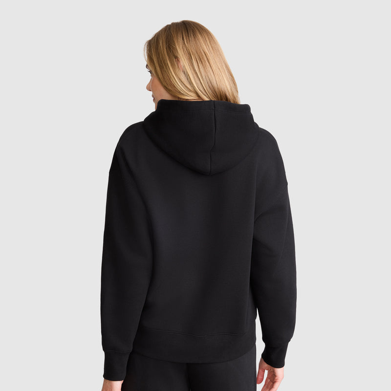 A person with light brown hair is shown from the back, wearing a Champion Tonal Script Freshman Powerblend Hoodie and black pants. The midweight fleece sweatshirt stands out against the light gray background.
