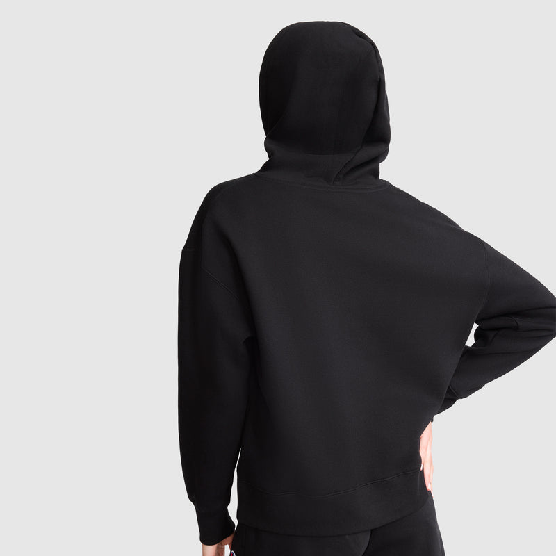 A person wearing a Champion Tonal Script Freshman Powerblend Hoodie and black pants stands with their back to the camera, one hand on their hip. The midweight fleece hoodie’s hood is up against a plain light gray background.