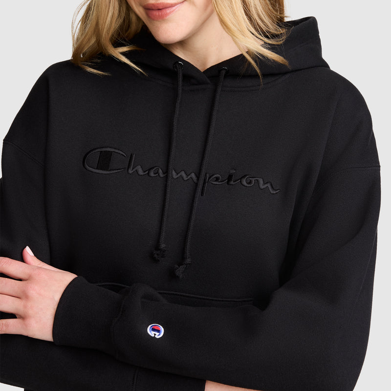 A woman stands with arms crossed against a plain background, wearing a black Champion Tonal Script Freshman Powerblend Hoodie featuring midweight fleece, an embroidered logo on the chest, and a small Champion patch near the wrist.