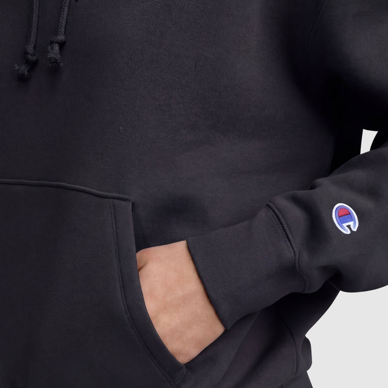 A person wearing a black Champion Tonal Script Freshman Powerblend Hoodie with one hand in the front pocket; the sleeve displays the Champion logo patch near the wrist.