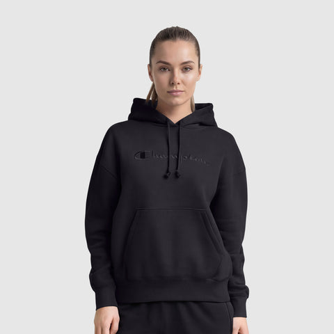 A woman with light hair pulled back wears a black Champion Tonal Script Freshman Powerblend Hoodie with a front pocket and embroidered logo, standing against a plain gray background.