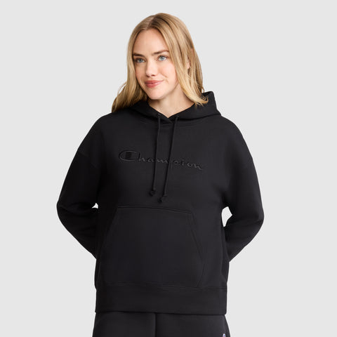 A woman with blonde hair wears the Champion Tonal Script Freshman Powerblend Hoodie, standing against a plain light gray background with her hands behind her back.