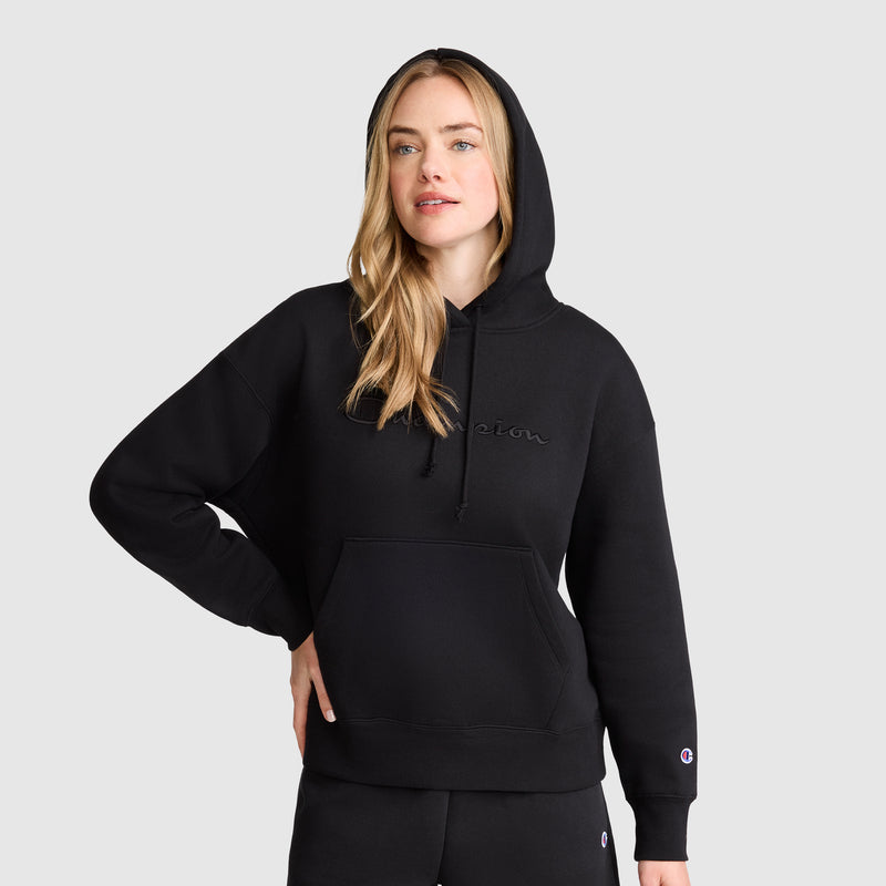 A woman with long blonde hair wears a black Champion Tonal Script Freshman Powerblend Hoodie and black sweatpants, standing against a light gray background with the midweight fleece hood up and one hand on her hip.