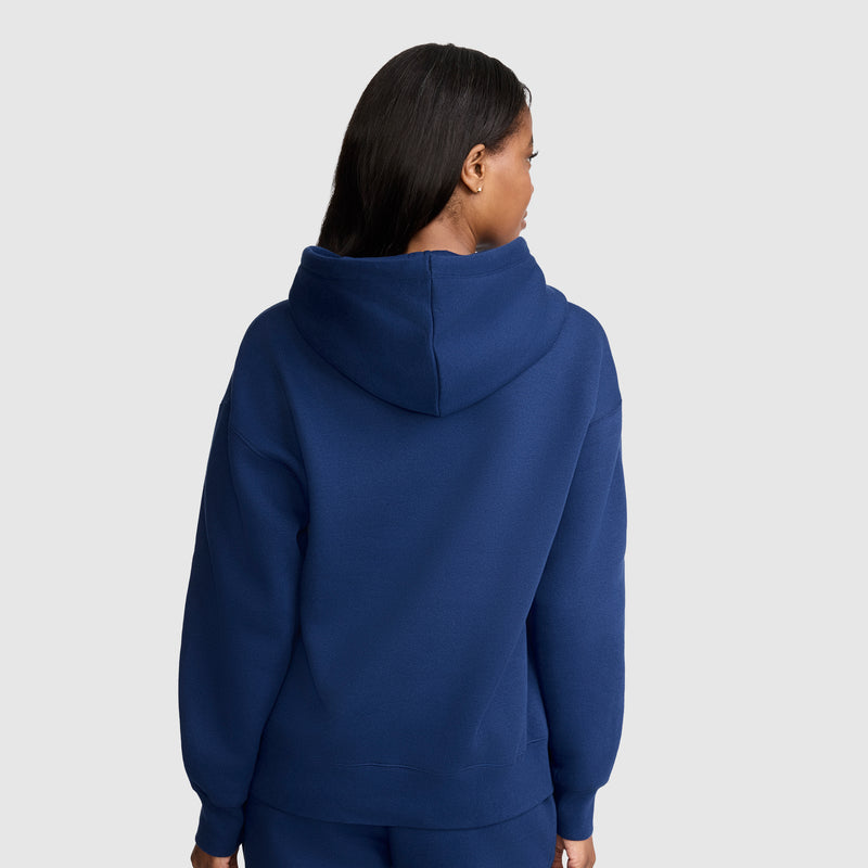 A person with long, dark hair is wearing a blue Champion Tonal Script Freshman Powerblend Hoodie and matching pants, standing with their back to the camera against a plain light gray background. This hoodie, crafted by Champion from soft midweight fleece, provides both comfort and style.