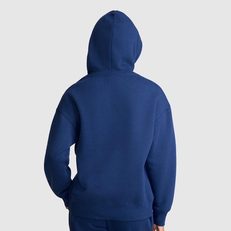 A person wearing a Champion Tonal Script Freshman Powerblend Hoodie, crafted from midweight fleece, stands with their back facing the camera, hands resting in the front pocket, against a plain light gray background.