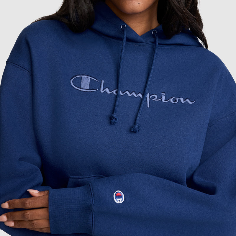 A person wearing a blue Champion Tonal Script Freshman Powerblend Hoodie, made from midweight fleece and featuring the embroidered logo on the chest and sleeve, stands with arms crossed.