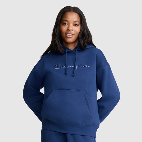 A woman wearing a blue Champion Tonal Script Freshman Powerblend Hoodie with her hands in the front pocket, smiling and standing against a plain light gray background. This midweight fleece hoodie from Champion blends comfort and style for a cozy, everyday look.