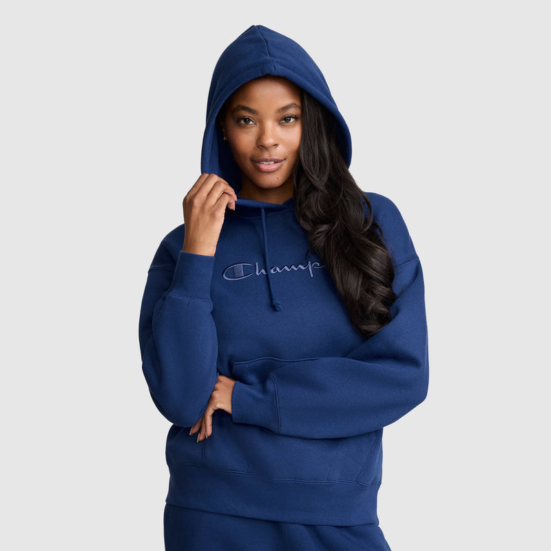 A woman wearing a blue Champion Tonal Script Freshman Powerblend Hoodie, crafted from soft midweight fleece, poses with one hand holding the hood and her other arm crossed at her waist against a plain light gray background.