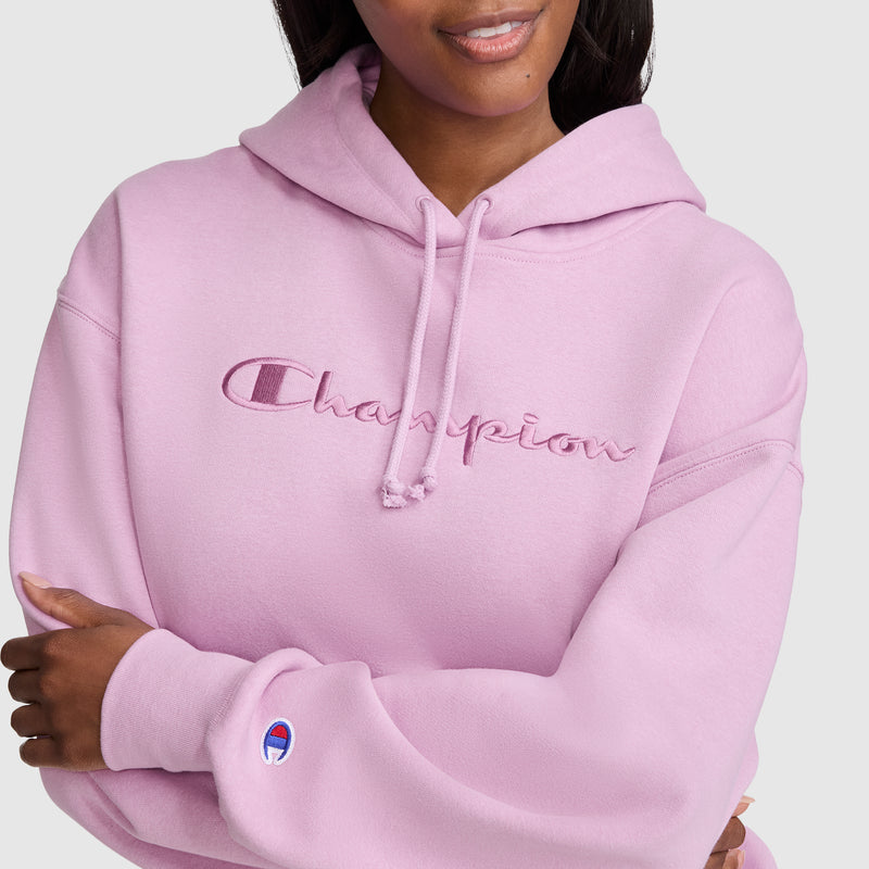 A woman wears a light pink Champion Tonal Script Freshman Powerblend Hoodie, featuring “Champion” embroidered across the front and the brand’s logo on the sleeve. This cozy fleece hoodie is styled as a women’s sweatshirt; she crosses her arms and smiles slightly.