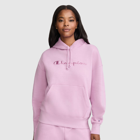 A woman wearing a light pink Champion Tonal Script Freshman Powerblend Hoodie stands against a plain light gray background, looking at the camera with a relaxed expression. This cozy sweatshirt features soft fleece fabric and the iconic Champion logo on the front.