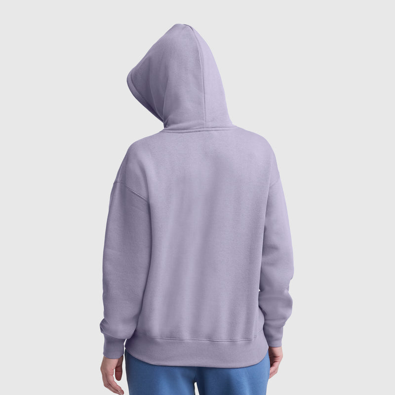 A person wearing a Champion Tonal Script Freshman Powerblend Hoodie in lavender, hood up, with blue pants, faces away from the camera against a plain light background.