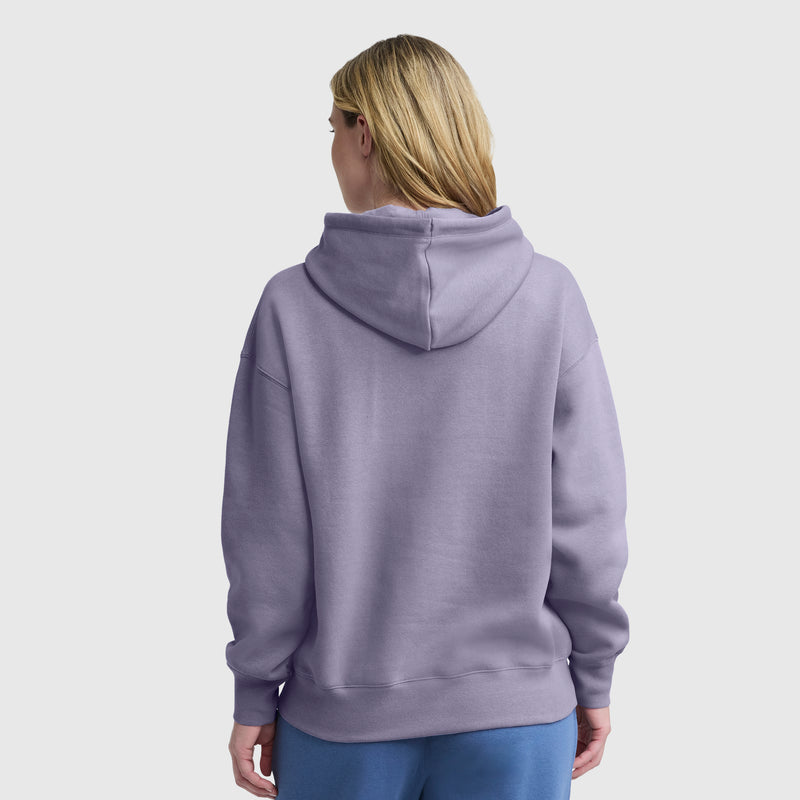 A person with long blond hair is seen from the back, wearing a light purple Champion Tonal Script Freshman Powerblend Hoodie and blue pants against a plain, light background.