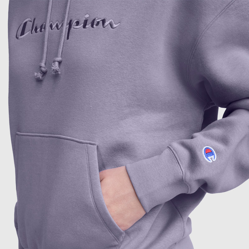 A person wears a lavender Champion Tonal Script Freshman Powerblend Hoodie with one hand in the front pocket. The women's pullover features an embroidered Champion logo on the chest and a small C logo on the sleeve.