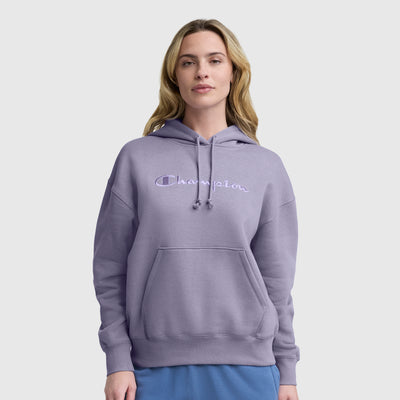 A woman with long, light brown hair wears a light purple Champion Tonal Script Freshman Powerblend Hoodie and blue pants, standing against a plain light gray background.