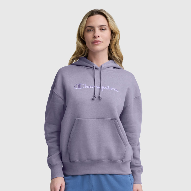 A woman with long, light brown hair wears a light purple Champion Tonal Script Freshman Powerblend Hoodie and blue pants, standing against a plain light gray background.