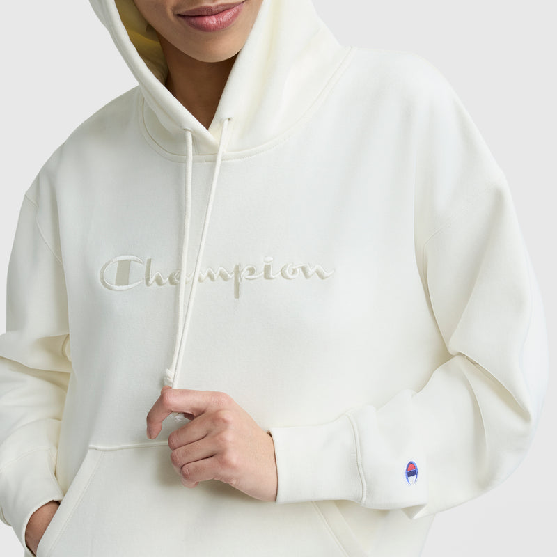 A person wearing a Champion Tonal Script Freshman Powerblend Hoodie in off-white, featuring an embroidered logo on the chest and a small sleeve patch, holds the drawstring while smiling slightly.