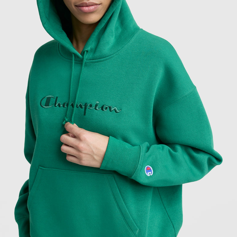 A person wears a green Champion Tonal Script Freshman Powerblend Hoodie with embroidered chest logo and sleeve patch, holding a drawstring. The background is plain light gray.