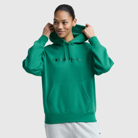 A person wearing a green Champion Tonal Script Freshman Powerblend Hoodie and light gray sweatpants smiles while adjusting the hood, standing against a plain light gray background.