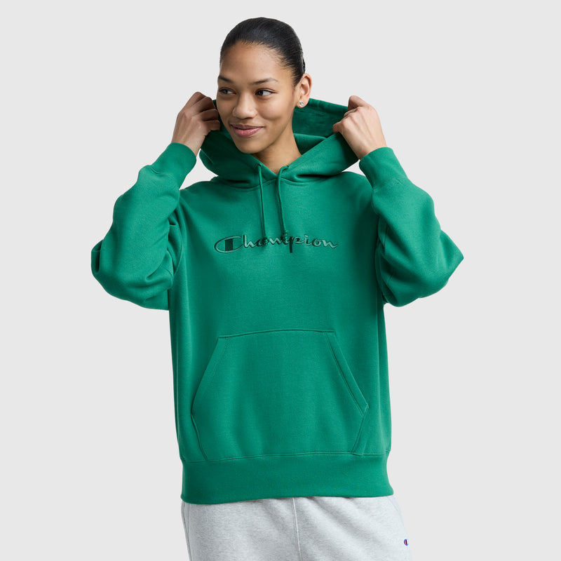 A person wearing a green Champion Tonal Script Freshman Powerblend Hoodie and light gray sweatpants smiles while adjusting the hood, standing against a plain light gray background.