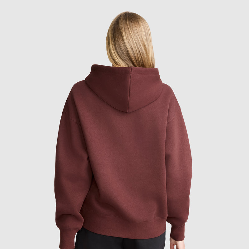 A person with straight, blonde hair is standing with their back to the camera, wearing a maroon Champion Tonal Script Freshman Powerblend Hoodie and black pants against a plain, light gray background.