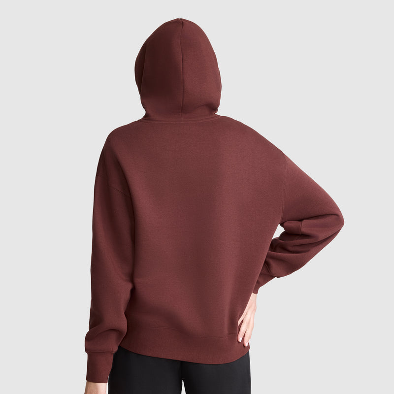 A person is shown from the back wearing a maroon Champion Tonal Script Freshman Powerblend Hoodie with the hood up. One hand rests on their hip, and they are paired with black pants. The Powerblend fleece offers comfort and warmth against a light gray background.