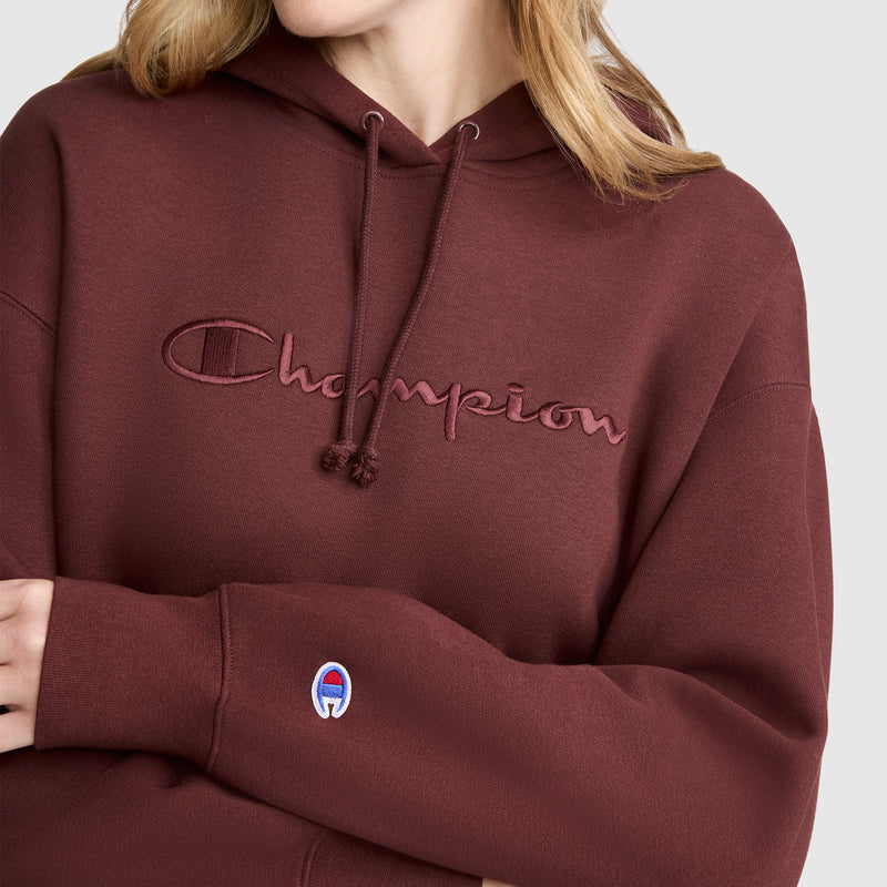 A person wears the Champion Tonal Script Freshman Powerblend Hoodie in maroon, featuring soft fleece fabric, an embroidered tonal script logo on the chest, and a small Champion emblem on the sleeve. Only their lower face, neck, and upper body are visible.