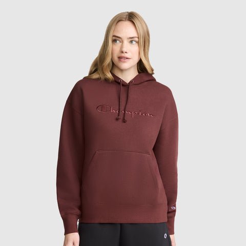 A woman stands against a plain light gray background, wearing the Champion Tonal Script Freshman Powerblend Hoodie in maroon—crafted from cozy fleece with a front pocket—and paired with black pants.