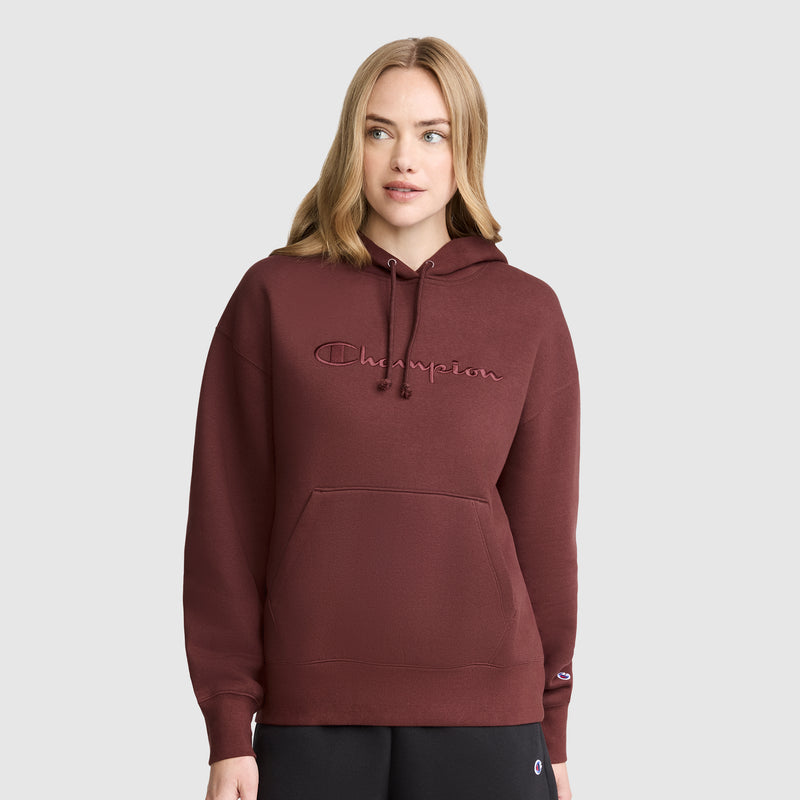 A woman stands against a plain light gray background, wearing the Champion Tonal Script Freshman Powerblend Hoodie in maroon—crafted from cozy fleece with a front pocket—and paired with black pants.