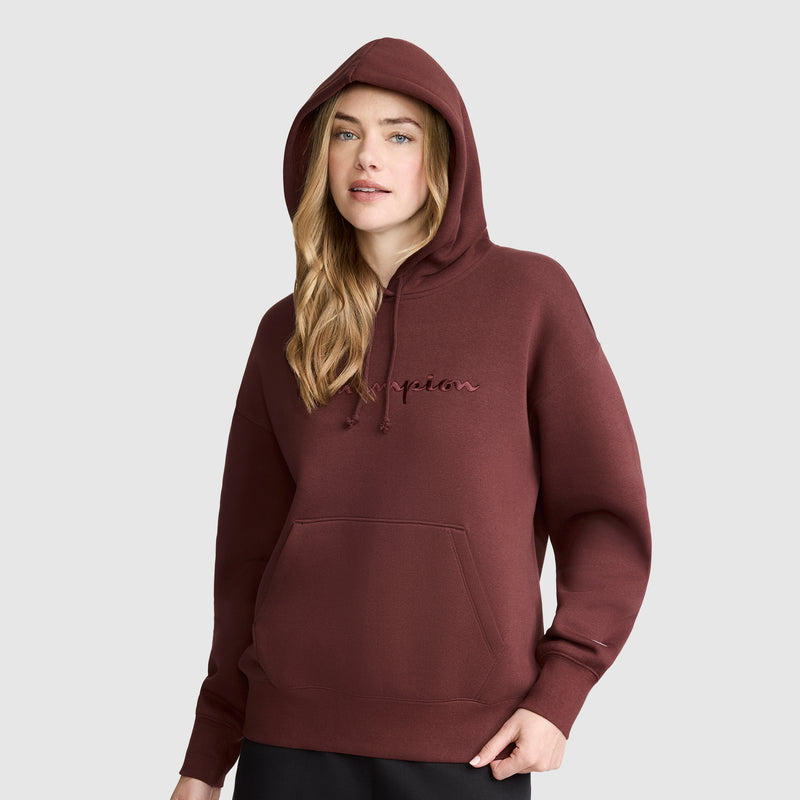 A woman wearing a maroon Champion Tonal Script Freshman Powerblend Hoodie, crafted from soft fleece and featuring a front pocket and signature logo, stands against a plain light gray background.