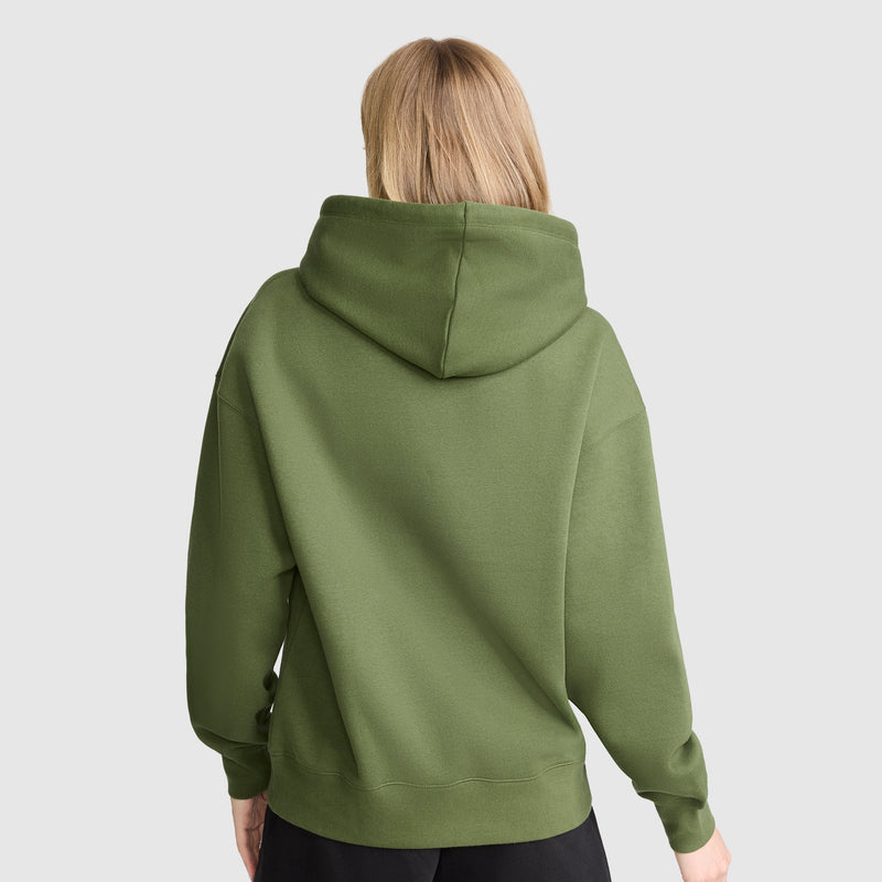 A person with blonde hair is shown from the back, wearing a Champion Tonal Script Freshman Powerblend Hoodie in olive green and black pants, standing against a plain light gray background.